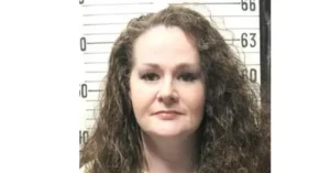 The image appears to be a mugshot of a woman with long, curly hair, taken against a height chart background.