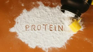 The image shows a close-up of a protein powder spill with the word "PROTEIN" written in the powder, accompanied by a yellow scoop and a black shaker bottle.