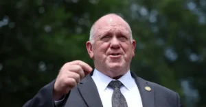 The image shows a man in a suit and tie, pointing with his right hand, set against a blurred background of greenery.