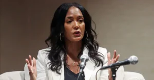 A woman is speaking into a microphone, gesturing with her hands as she addresses an audience.