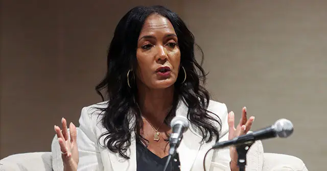 A woman is speaking into a microphone, gesturing with her hands as she addresses an audience.