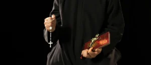 The image shows a person in a black robe holding a rosary in one hand and a book with a crucifix on the cover in the other.