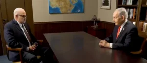 Two men in formal attire are seated across from each other at a table in a room with bookshelves and a map on the wall.