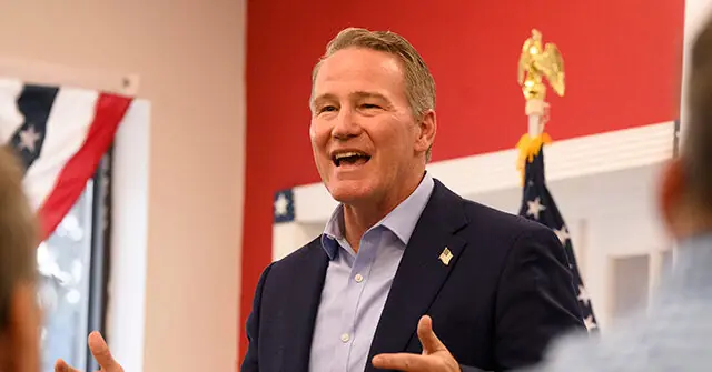 The image shows a man in a suit smiling and gesturing, with an American flag and a decorative eagle in the background.