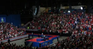 The image depicts a large rally with a stage and a crowd of people, many wearing red, in a venue adorned with patriotic decorations.