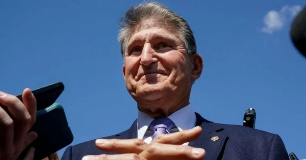A man in a suit and tie is speaking outdoors under a clear blue sky, with microphones and a smartphone visible in the foreground.