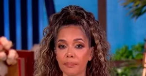 The image shows a person with curly hair, wearing earrings, in what appears to be a studio setting with a blue background and some greenery.