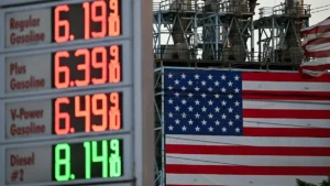 The photo shows a gas price sign displaying high fuel costs alongside an American flag and industrial equipment, symbolizing the economic impact of rising fuel prices.
