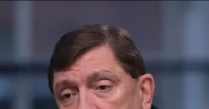 The image shows a close-up of a person with short brown hair, wearing a dark suit, against a blurred background.