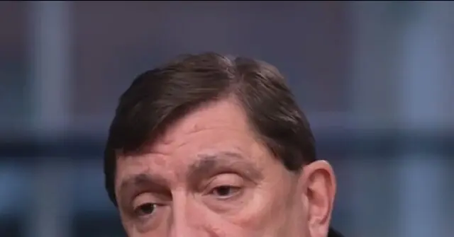 The image shows a close-up of a person with short brown hair, wearing a dark suit, against a blurred background.