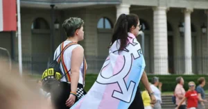 Two individuals walk together, one draped in a transgender flag, symbolizing support and pride for the LGBTQ+ community.