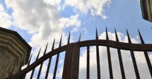 The image shows a spiked metal fence against a backdrop of a partly cloudy sky.