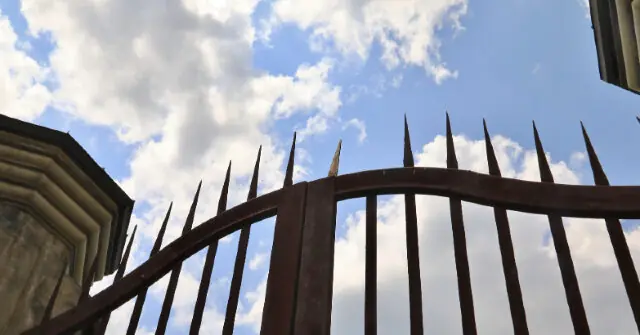 The image shows a spiked metal fence against a backdrop of a partly cloudy sky.