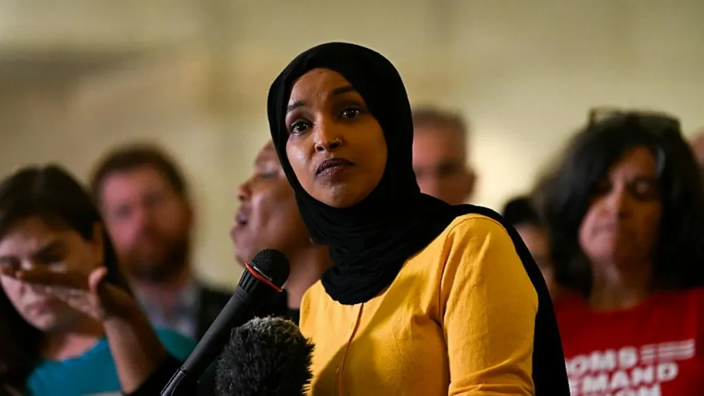 A woman wearing a hijab speaks into a microphone at a public event, with an audience in the background.