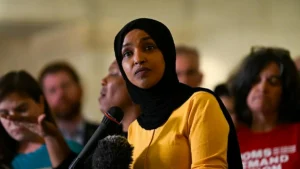 A woman wearing a hijab speaks into a microphone at a public event, with an audience in the background.