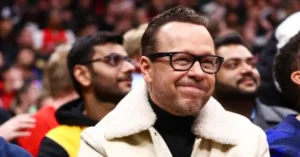 The image shows a man in a crowd, wearing a white jacket with a fur collar and a black turtleneck, smiling and looking to the side.