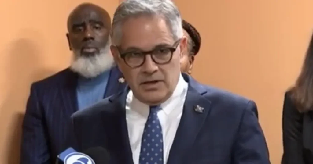 A man in a suit and tie is speaking at a press conference, with other individuals standing behind him.