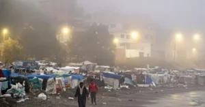 The photo depicts a foggy scene of a makeshift camp with tents and debris, illuminated by streetlights, with two individuals walking in the foreground.