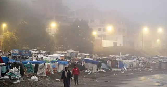 The photo depicts a foggy scene of a makeshift camp with tents and debris, illuminated by streetlights, with two individuals walking in the foreground.