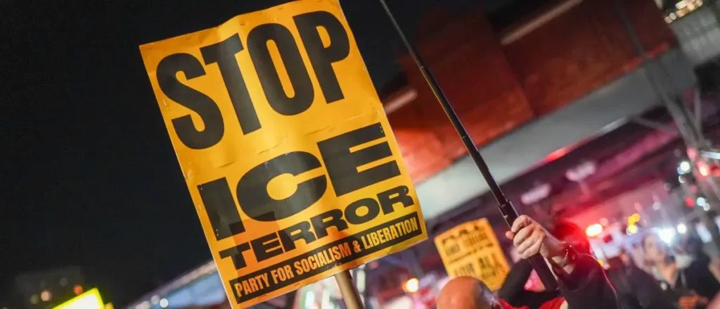 The photo depicts a protest sign reading "STOP ICE TERROR" held up at a nighttime demonstration.