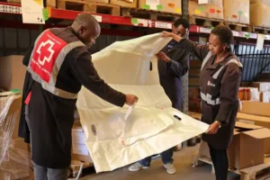 Three individuals in Red Cross uniforms are working together to unpack and organize supplies in a warehouse.