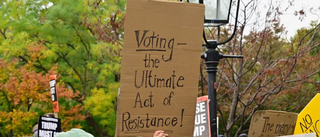 The photo shows a protest sign that reads, "Voting – the Ultimate Act of Resistance!" with trees and other signs in the background.