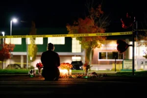 A person kneels at a makeshift memorial with candles and flowers, surrounded by police tape, at night.