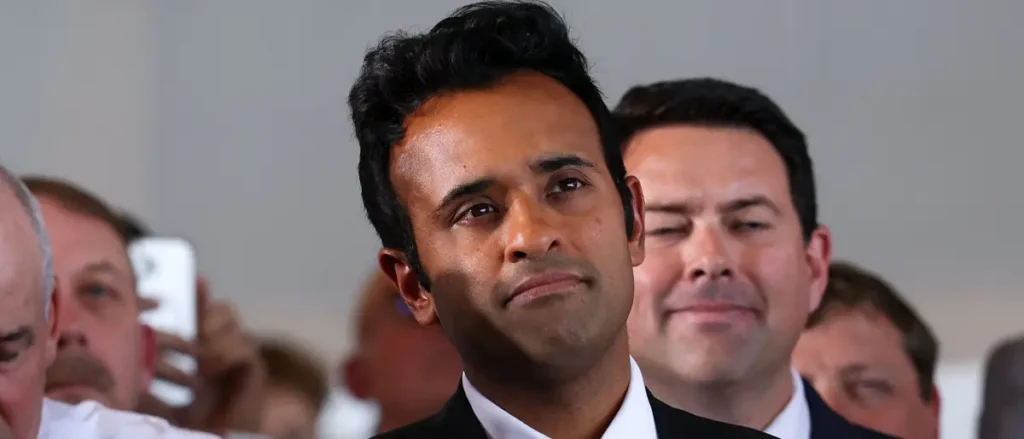 The photo shows a man in a suit standing in a crowd, with another man behind him smiling.