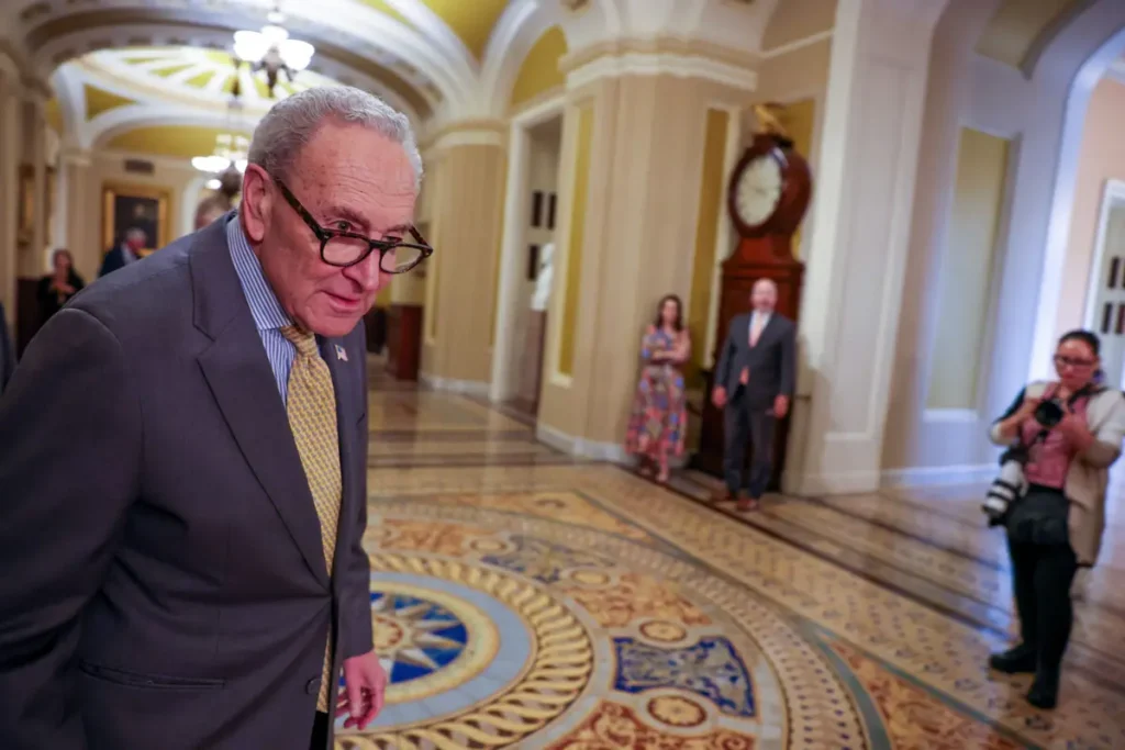 A man in a suit and tie walks through a grand, ornate hallway, with other individuals in the background.