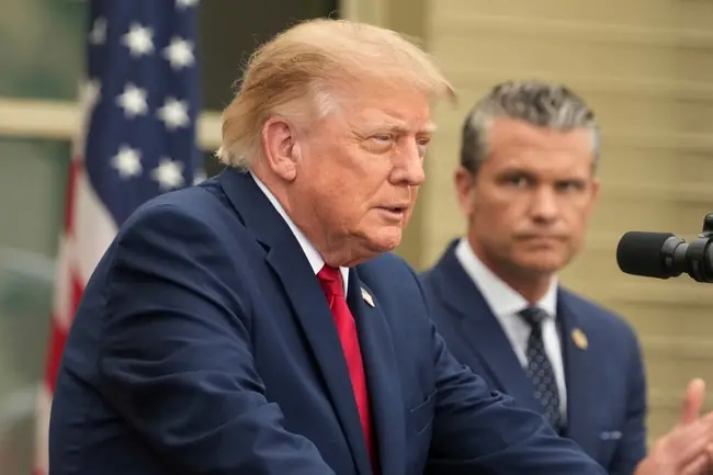 The image shows a man in a suit speaking at a podium with an American flag in the background.