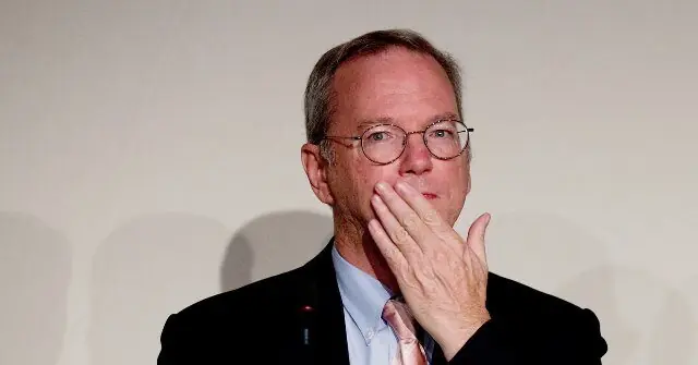 The image shows a man in a suit and tie, wearing glasses, with his hand near his mouth, possibly in a thoughtful or contemplative pose.