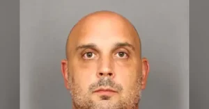 The image is a mugshot of a bald man with a short beard and mustache, taken against a plain background.