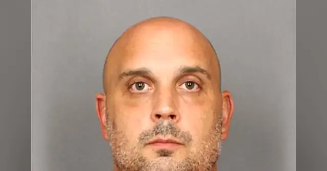 The image is a mugshot of a bald man with a short beard and mustache, taken against a plain background.