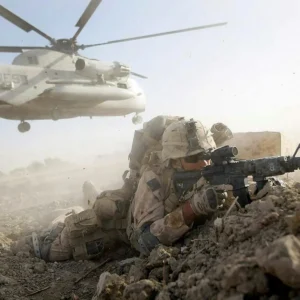 A soldier takes cover while a helicopter hovers in the background during a military operation.
