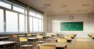 The image shows an empty classroom with rows of desks and chairs, a chalkboard, and a clock on the wall.