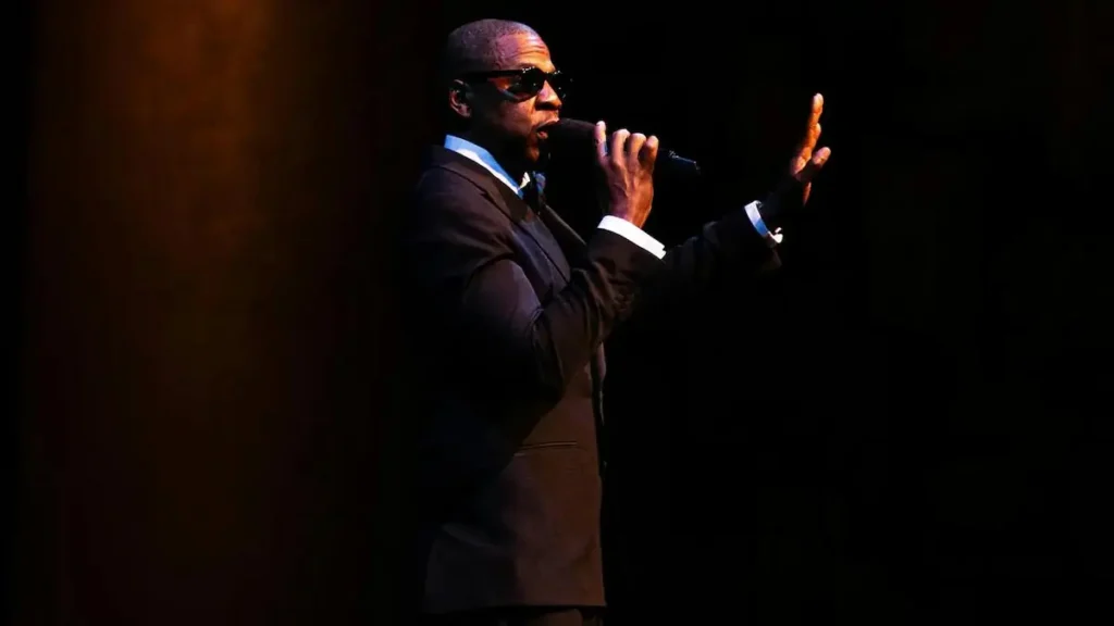 A man in a suit and sunglasses is performing on stage, holding a microphone and gesturing with his other hand.
