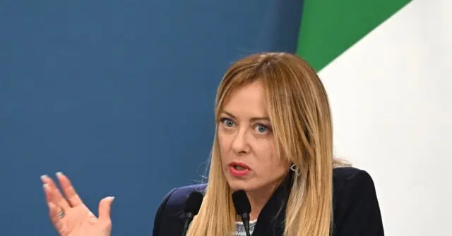 A woman with long blonde hair is speaking into a microphone, gesturing with her hand, in front of a blue and green background.