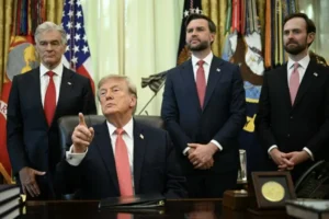 The image shows a group of men in formal attire, including one seated at a desk, in what appears to be a formal government setting.