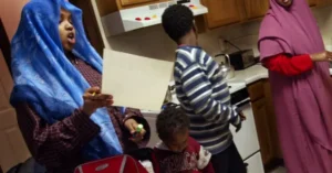 A family is gathered in a kitchen, with one person holding a piece of paper and another child looking at it, while others stand nearby.