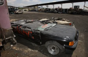 The image shows a damaged and partially dismantled car in a junkyard, surrounded by other wrecked vehicles.