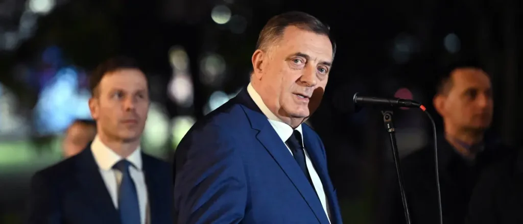 A man in a blue suit stands at a podium, speaking into a microphone, with two other individuals in the background.