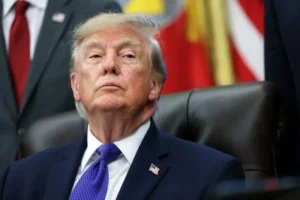 The image shows a man in a suit and tie, seated in front of an American flag, appearing to be in a formal setting.