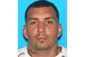 The image is a mugshot of a man with short dark hair, a goatee, and visible tattoos on his neck and chest.