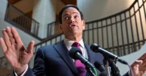 A man in a suit and tie is speaking at a press conference, gesturing with his hands.