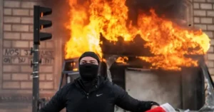 A person wearing a black mask and jacket stands in front of a burning building with graffiti on the wall.