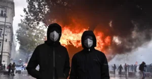 The image depicts two individuals wearing masks and hooded jackets standing in front of a large fire and smoke, with a crowd of people in the background, suggesting a protest or demonstration.