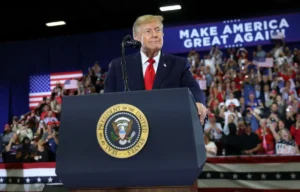 The image shows a political rally with a speaker at a podium featuring the presidential seal, addressing a crowd with a "Make America Great Again" backdrop.