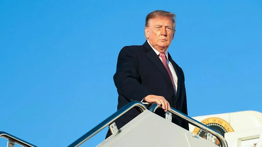 The image shows a person in a suit standing on the steps of an aircraft, with a clear blue sky in the background.