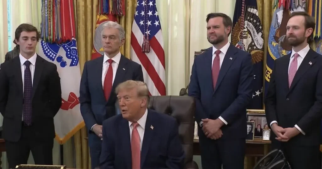 The image shows a formal meeting in the Oval Office with a seated individual and four standing men in suits, with American flags and presidential seals in the background.
