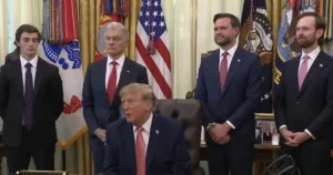 The image shows a formal meeting in the Oval Office with a seated individual and four standing men in suits, with American flags and presidential seals in the background.